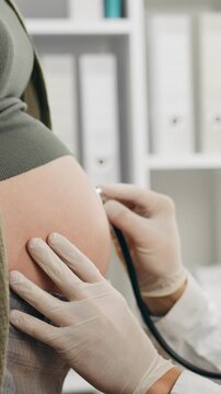 Doctor in white coat conducts a detailed abdominal examination of a pregnant patient in a hospital waiting area, using a stethoscope to monitor fetal movements as part of routine prenatal assessment