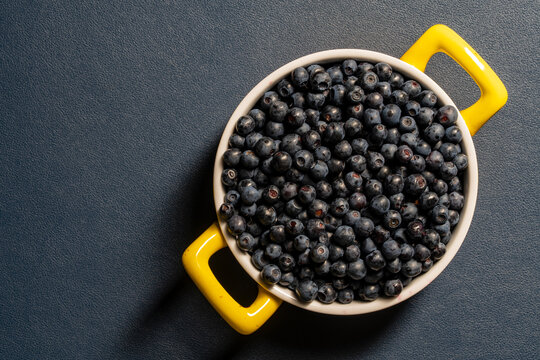 Fresh blueberries in white bowl with yellow handles. Top view round bowl filled with ripe wild blueberries on blue textured background with copy space representing healthy food and summer harvest
