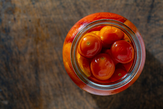 Pickled cherry tomatoes in open glass jar. Top view of marinated red and yellow cherry tomatoes inside an open glass jar on rustic wooden table background with copy space representing homemade food