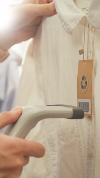 In a clothing store, a manager points a red-laser barcode scanner at apparel tags, reading each code, while an Asian worker stands nearby handling inventory paperwork, as part of stock accounting