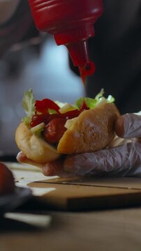 Inside the industrial kitchen of a street diner, a hot dog maker squeezes bright red tomato ketchup over a sausage nestled in a bun, emphasizing fast-food preparation and the routine service flow