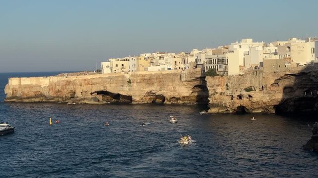 Small boats cruising along the limestone coast of Polignano a Mare, Puglia, Italy on the calm Adriatic Sea with historic waterfront in warm evening light. Summer travel, tourism, vacation, coastal lan
