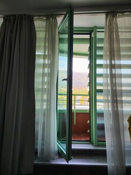 Open green framed window with white sheer curtains in dark room interior, mountain and city view visible through open window, balcony railing visible, daylight outside