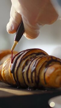 Pastry chef wearing uniform drizzles melted chocolate over freshly baked croissant on a stainless-steel work surface in a bakery, the glossy coating spreading as part of the pastry finishing process