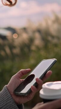 Woman stands on an urban street sidewalk, holding a disposable paper coffee cup, taking a sip while glancing at her phone, bright sunlight pours over the scene, emphasizing a typical sunny-day pause