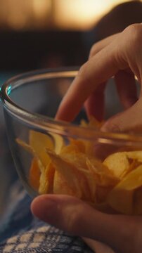 Young couple sits on plush sofa in dimly lit home theater, sharing bowl of potato chips while a TV screen glows with a series, the couple appears comfortable, legs tucked, creating a calm atmosphere