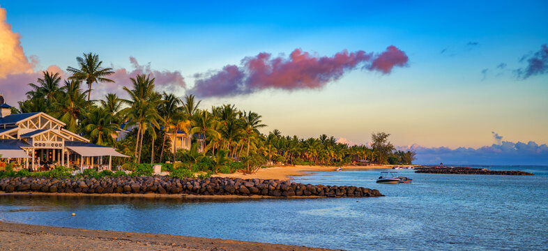 Panoramic sunset view of a tropical beach in Bel Ombre, Mauritius, with palm trees, calm lagoon water, luxury coastal resorts and colorful sky.