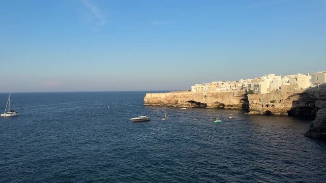 Wide coastal view of the historic cliffside old town in Polignano a Mare, Puglia, Italy overlooking the Adriatic Sea with boats and limestone cliffs in warm evening light. Summer travel, tourism, vaca