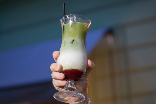a hand holding a glass of layered iced matcha latte with ice cubes and a black straw raised into the air against a blurred background