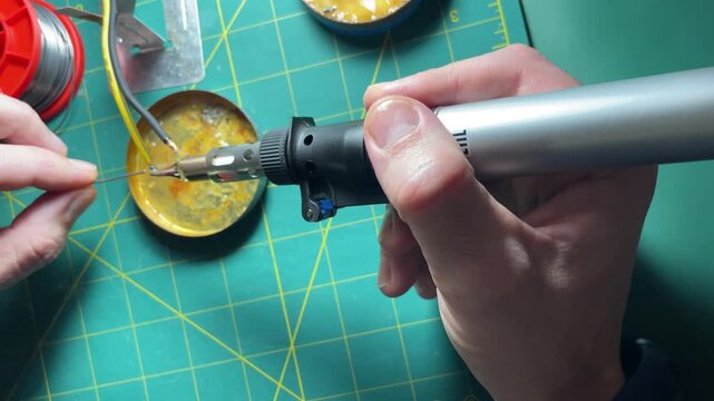 Close up of worker hands using portable cordless gas soldering iron torch to melt rosin and wire with smoke on green cutting mat backdrop at workplace. High quality 4k footage