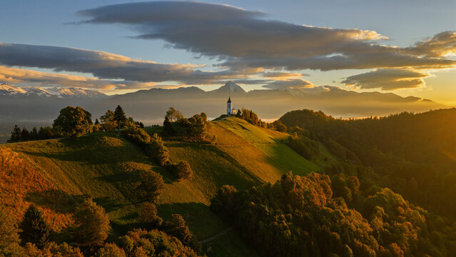 Aerial view of the Church of Saints Primus and Felician on a hilltop surrounded by rolling green hills and autumn forests with a mountain backdrop in Jamnik, Kranj, Slovenia.