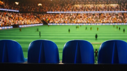 Close-up of stadium seating facing blurred soccer field with players waiting for ball kickoff during match day, empty blue seats inside sports venue and athletes competing in football tournament.