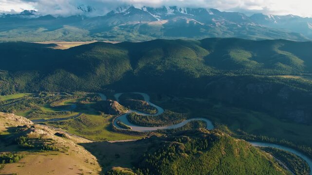 Aerial slow tracking shot of winding river meandering through Altai mountainous valley and forested foothills at golden morning light.