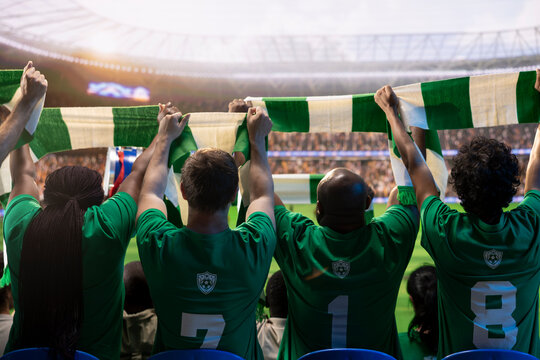 Rear view of football supporters in sports jerseys raising hands with scarves in packed stadium, cheering crowd holding match day merchandise.