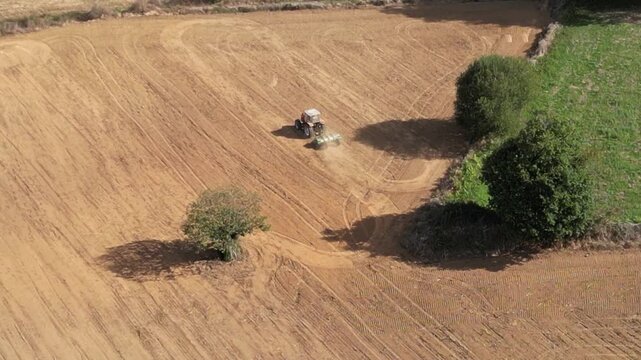 An aerial footage of a tractor working dry farmland along the Camino de Santiago in Spain, surrounded by scattered trees and warm daylight