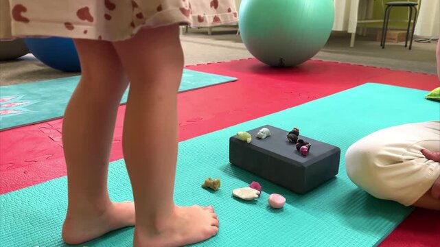 A girl with flat feet is engaged in children's rehabilitation, lifts toys to the black step. Green rubber mat. Treatment of clubfoot in a child