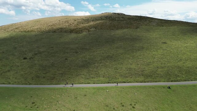 An aerial view of three figures walking on a paved road ,cutting across the base of a sloping green hillside on the Camino de Santiago trail in France