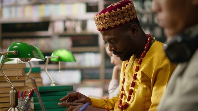 African mature student in yellow traditional clothing smiles while working on laptop at desk in study area with books, monitors and classmates nearby.