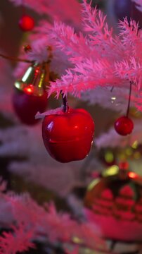 Red apple christmas ornament hanging on white tree with colorful lights and golden bells bokeh background vertical video footage.