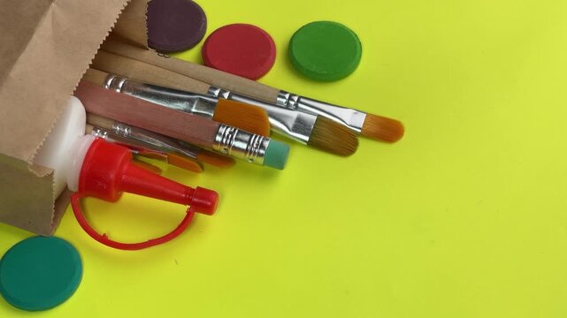 Art supplies spilling from paper bag panning video footage paintbrushes glue bottle and watercolor discs on yellow background with copyspace. 