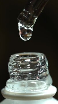Dropper releasing clear serum drop over glass cosmetic jar macro closeup on black background luxury skincare and beauty concept. 