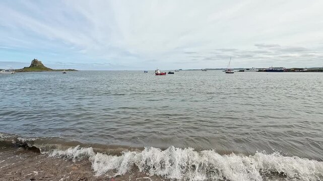 A slow-motion push-in shot of boats moored off Holy Island on the North East coast, gradually closing in on calm water, anchored vessels and the rugged shoreline.
