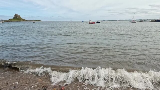 A slow-motion panning shot of boats moored off Holy Island on the North East coast, sweeping across calm waters, anchored vessels, and the rugged shoreline backdrop.