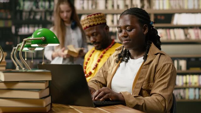 Woman learner studying on laptop and typing beside books, many textbooks on shared wooden table in library, classmates and bookshelves blurred in background, for education technology.