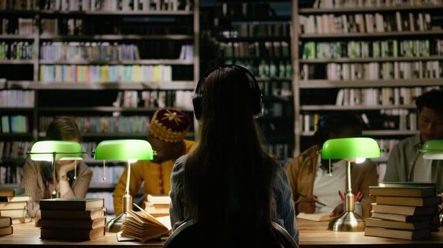 Back view of student adjusting headphones in dark library study room, multitasking with focus music near green lamps and books, for education technology.