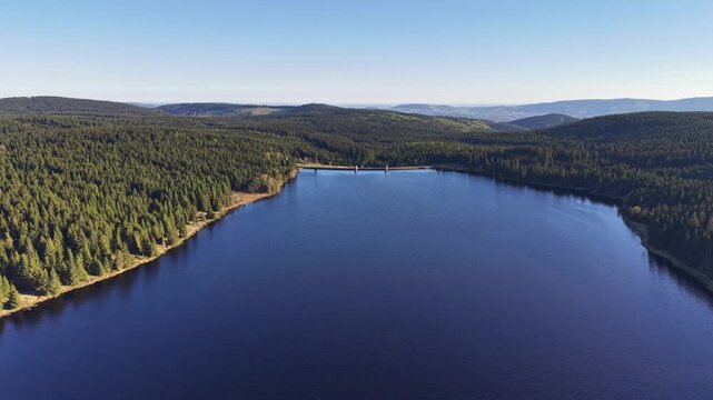Aerial reservoir lake with dam forested shoreline mirror water glassy surface pine slopes wide horizon calm