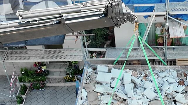 Construction workers using a crane to lift debris from a demolition site. Workers guide a crane lifting a large container filled with rubble and construction waste from a building demolition project.