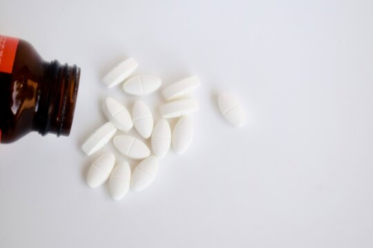 Top down view of white oblong prescription medicine caplets scattered on a white surface next to an open amber glass bottle.