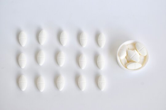 Top-down minimalist view of a 3x5 grid arrangement of white oblong tablets paired with an overflowing white bottle cap on a seamless white backdrop. Pharmaceutical auditing concept.