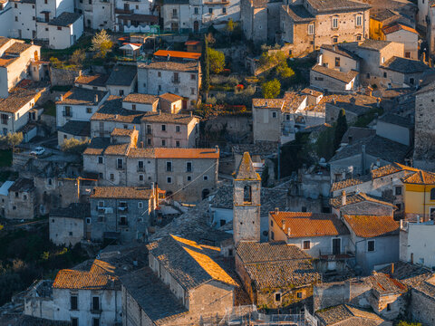 Aerial view of the historic town of Capestrano, featuring stone buildings with terracotta rooftops, a bell tower, and narrow streets bathed in golden hour light in Capestrano, Abruzzo, Italy.