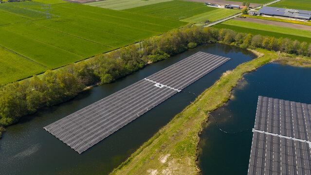 Aerial view of a large floating solar panel array installed on a calm pond, surrounded by lush green agricultural fields and trees in Zwolle, Overijssel, Netherlands.