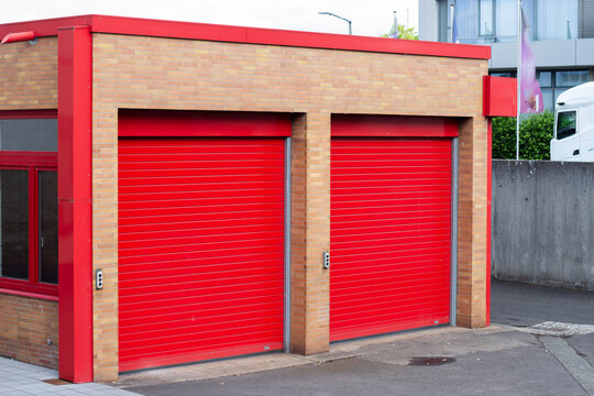 Red roller shutter doors on a brick building exterior with a concrete sidewalk and modern architecture visible in the background