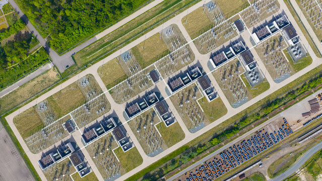 Aerial view of an electrical substation with rows of high-voltage transformers and switchgear on green grass, adjacent to a large parking lot in Velsen-Noord, North Holland, Netherlands.
