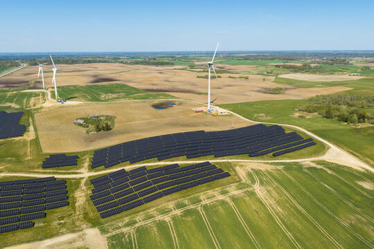 Aerial view of a solar farm with rows of photovoltaic panels and wind turbines in a rural landscape with green and brown fields in Jacionys, Vilnius County, Lithuania.