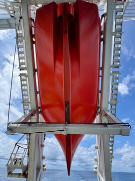 Low angle view of an enclosed rescue lifeboat suspended on davits aboard a cargo ship, showing maritime safety equipment against a bright blue sky with clouds