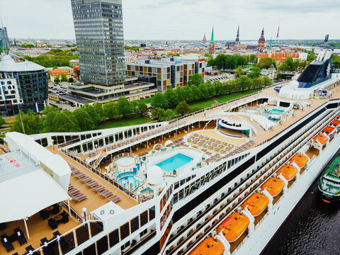 Aerial view of a large cruise ship docked in the river with a swimming pool, deck chairs, and lifeboats visible on the upper deck, set against the urban skyline of Riga, Latvia.