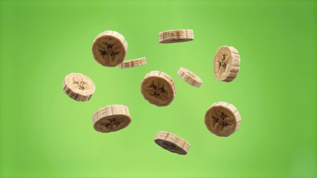 Multiple round cuts of ripe banana fruit floating and scattering through the air in a slow-motion effect. The clip includes a solid green screen backdrop designed for effortless post-production editin