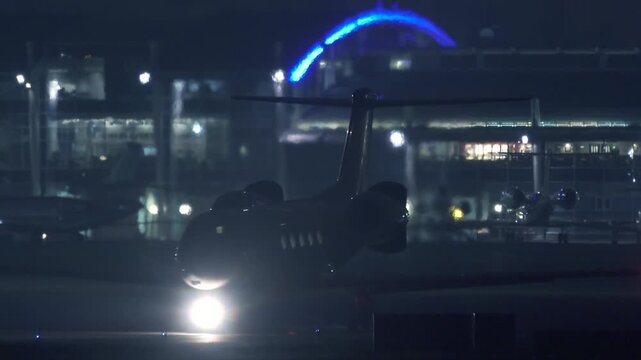 Private Business Jet Taxiing on Runway at Night Before Takeoff: Airport Terminal Lights and Parked Aircraft in Background | Aviation Ground Operations Telephoto Shot