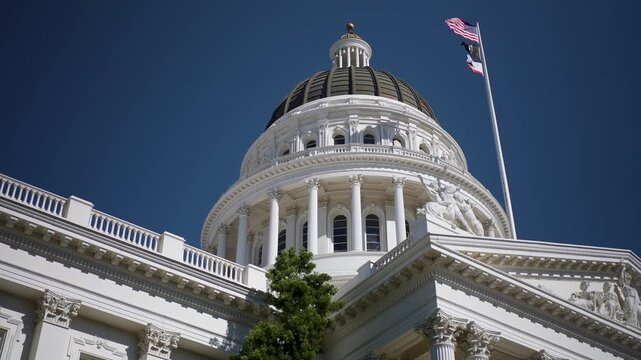 California State Capitol stands tall against blue sky with flags flying high and trees surrounding the area on sunny day
