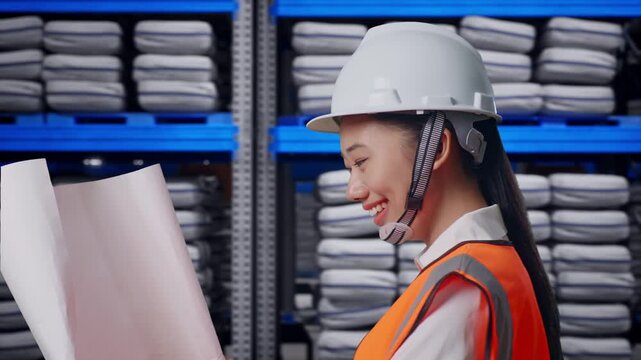 Side View of Asian Female Engineer Looking At Blueprint In His Hands While Walking at Warehouse storage with stacked white sacks