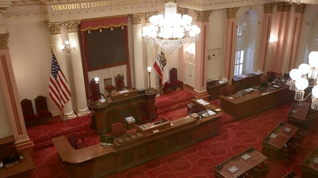 The video shows the interior of the California State Senate Chamber in Sacramento with desks, flags, and lights during a quiet time.