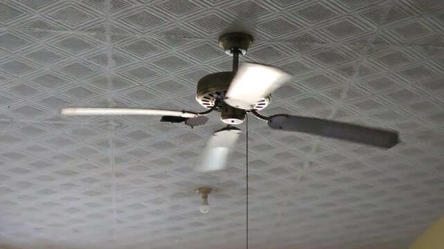 An old ceiling fan spinning on a patterned ceiling inside a home with a bare lightbulb in the background