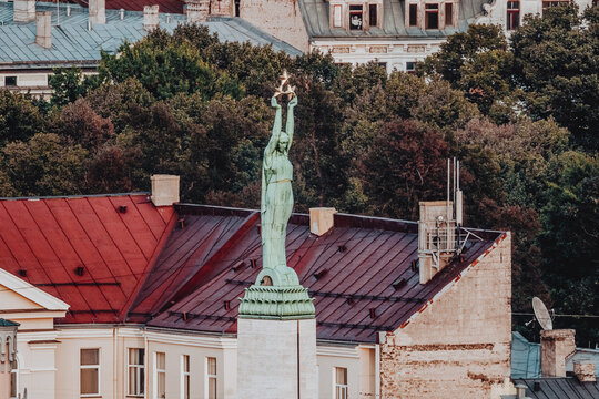 Aerial view of the Freedom Monument featuring its iconic copper statue atop a stone pedestal, surrounded by historic red metal rooftops and lush green trees in Riga, Latvia.