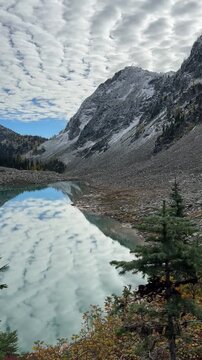 Beautiful alpine lake reflecting a patterned cloudy sky beneath rugged mountain peaks with a light dusting of snow