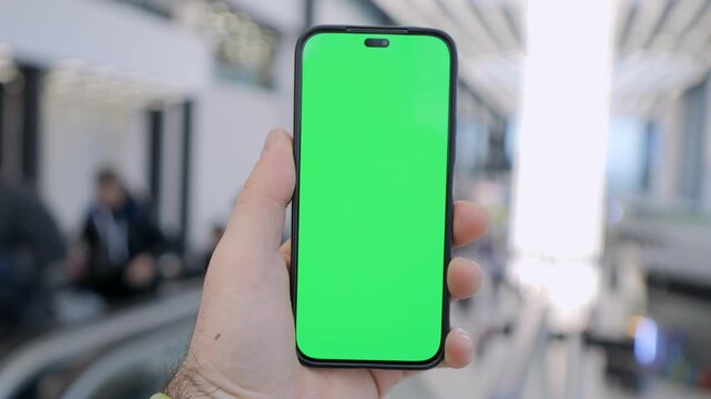 Hand holds smartphone with green screen. Background shows blurred indoor space. Person's emotion is neutral. Ideal for app previews or digital marketing. Perfect for editing with custom visuals