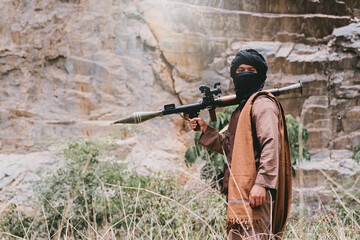 Fototapeta premium Outdoor military concept. Armed man in rocky canyon holding rocket launcher, masked face and desert clothing